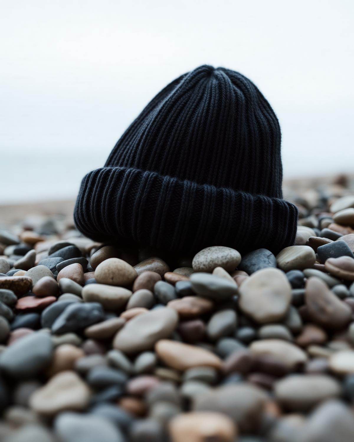 Brightonkelp charcoal ribbed merino wool beanie resting on smooth Brighton shingle pebbles